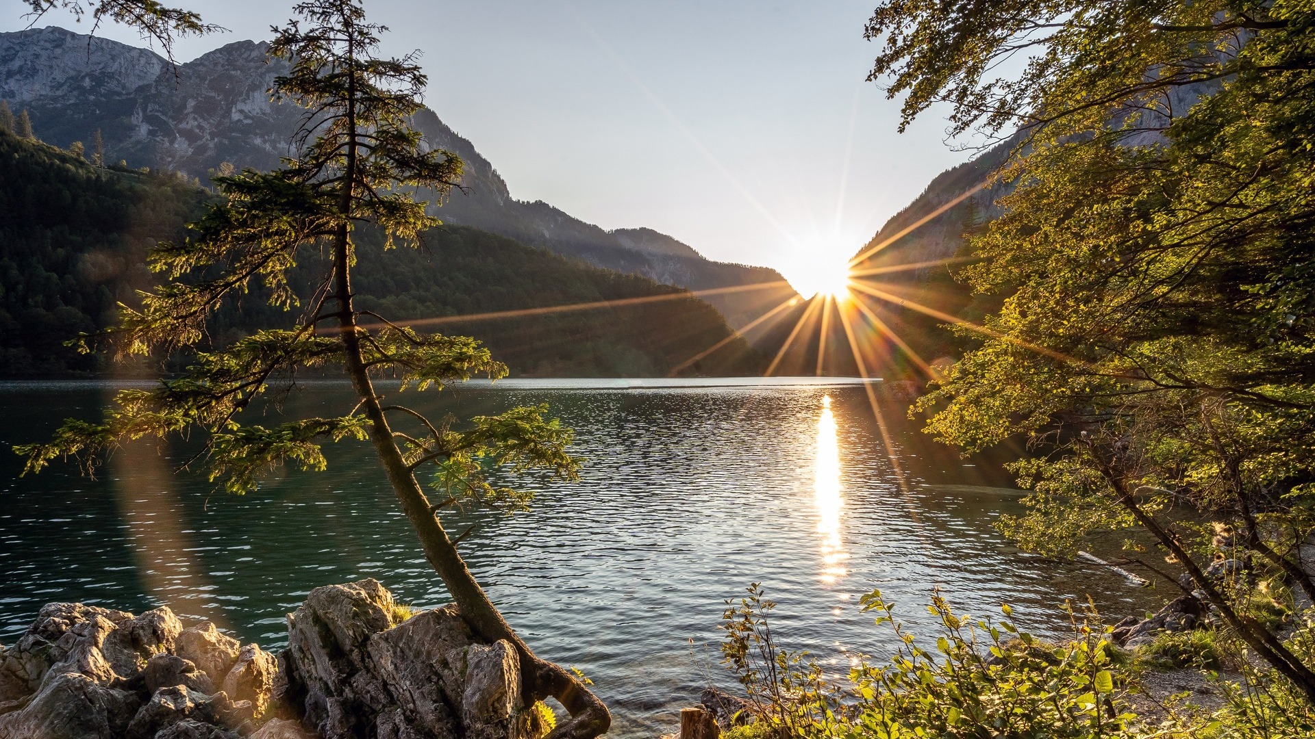 Calm mountain lake at sunrise with golden sun rays reflecting on the water between forested slopes and a rocky shoreline